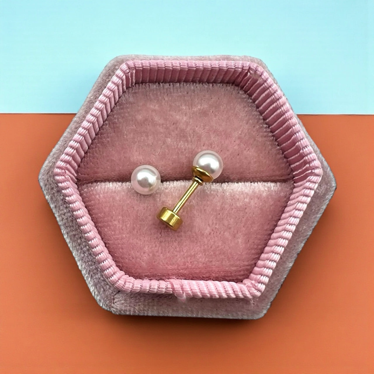 Pair of pearl earrings with gold posts in a pink hexagonal jewelry box on a two-tone background.
